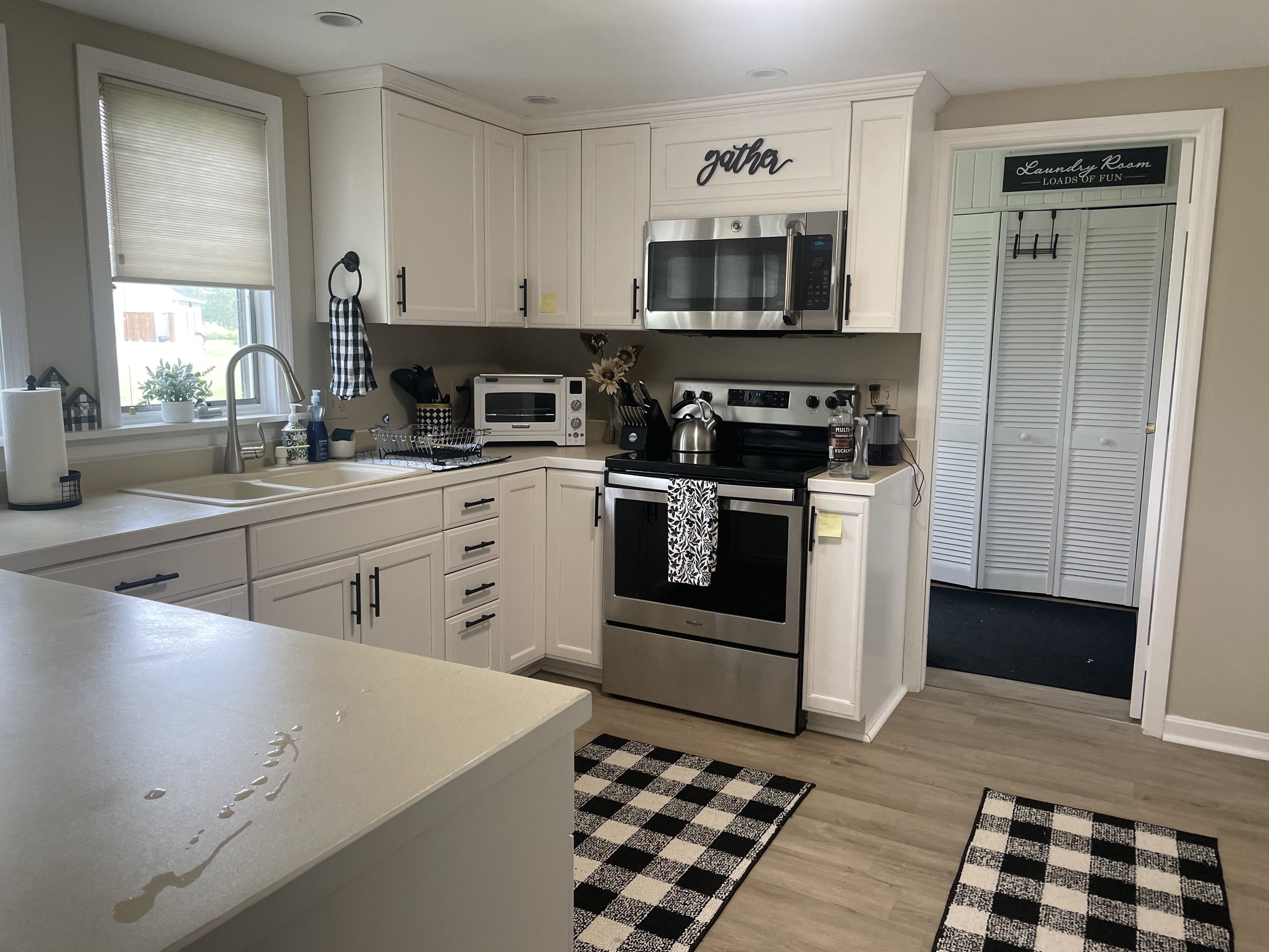 Bright white kitchen with stainless appliances, corner sink, and checkered floor rugs
