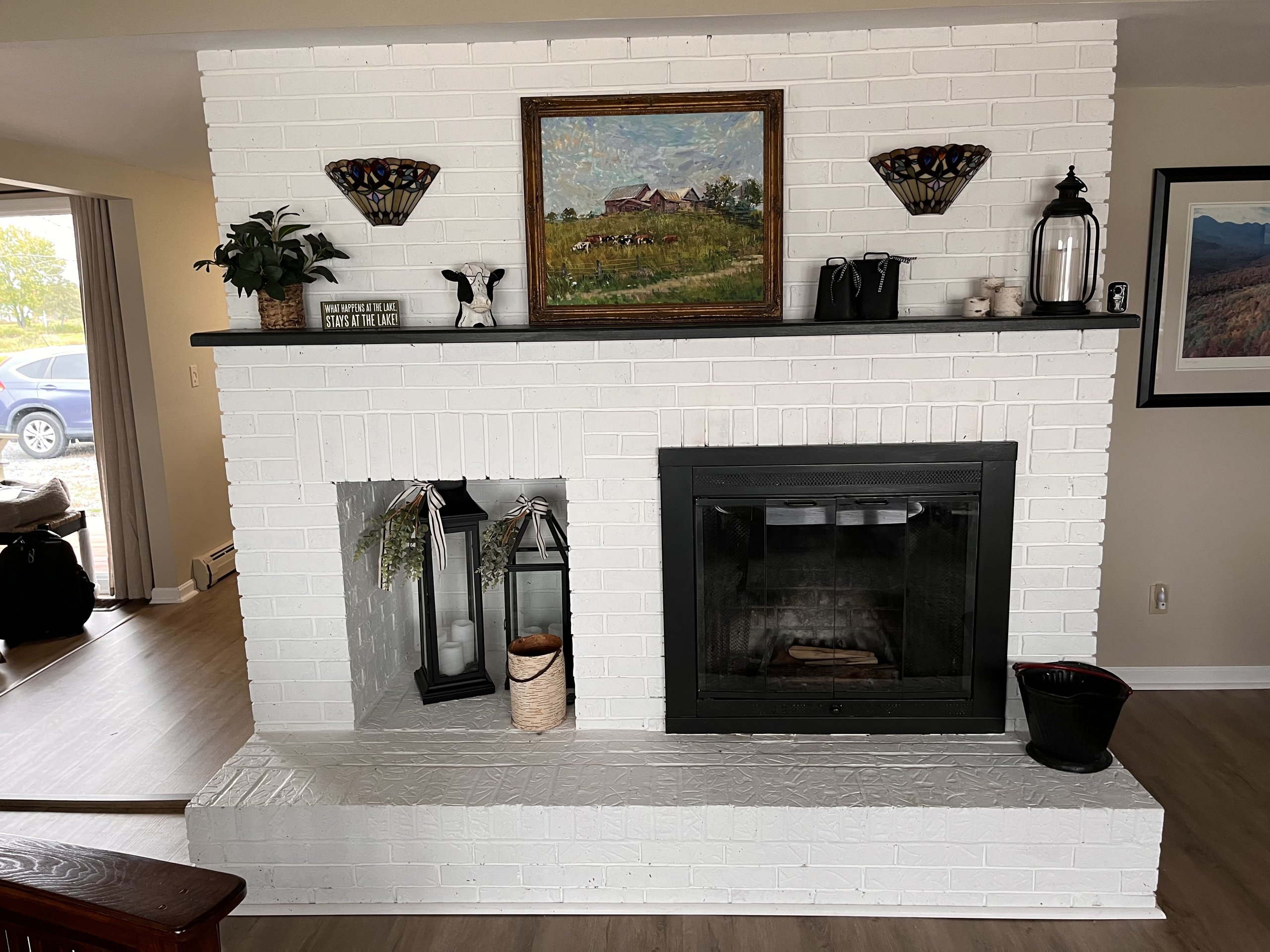 Fireplace mantel styled with a farm painting, plants, and lake-house accents