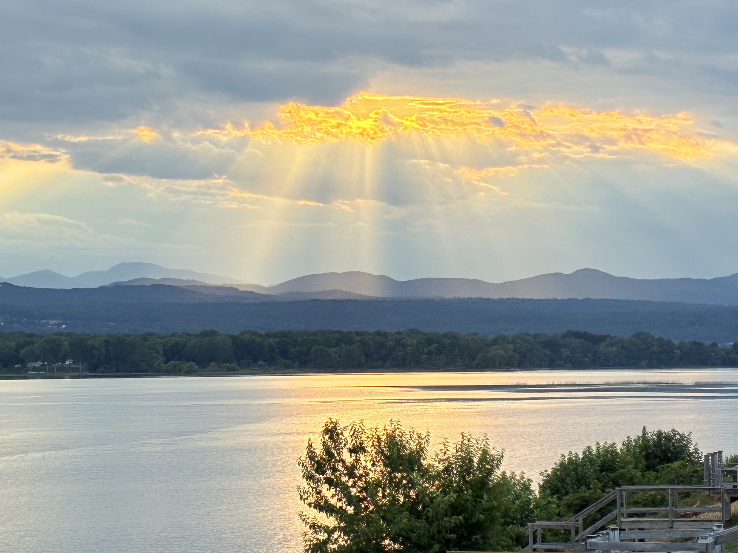 Evergreen branches frame a dramatic sunburst breaking through clouds over the lake